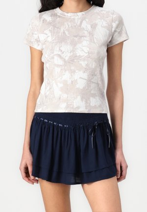 Femme portant un haut beige à manches courtes à motifs, rentré dans une mini-jupe à volants bleu marine avec taille à cordon.