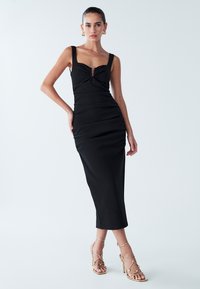 Vestido midi negro ajustado con escote cuadrado, detalles fruncidos y cierre delantero con cremallera. Combinado con sandalias doradas de tacón alto y tiras.