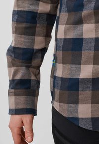 Personne portant une chemise en flanelle à carreaux bleue, marron et beige avec une petite étiquette du drapeau suédois sur le côté, et un pantalon noir visible en bas.