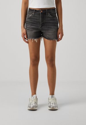 Kvinde iført sorte slidte denimshorts, hvid croppet top og hvide sneakers, der står mod en ensfarvet lysegrå baggrund.