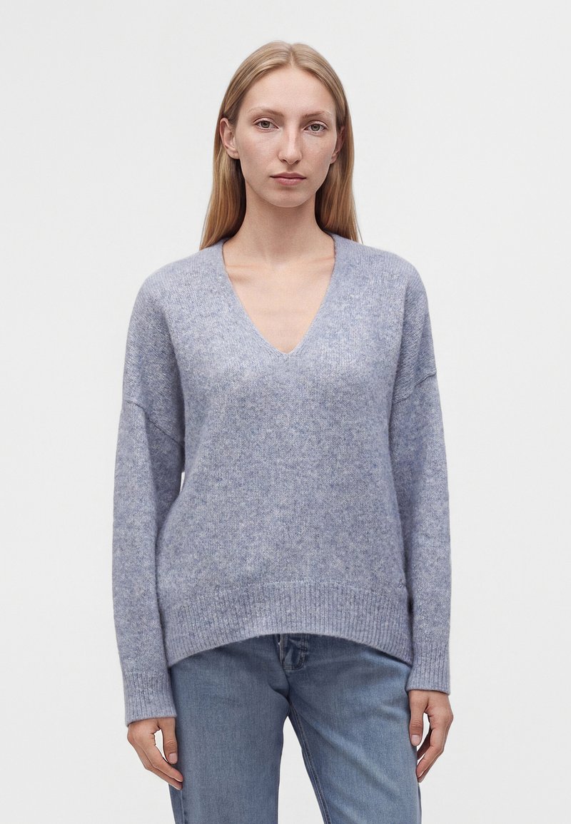 Jeune femme aux longs cheveux blonds portant un pull violet clair à col en V et un jean bleu, debout devant un fond blanc uni.