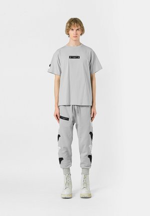 Camiseta de manga corta color gris claro con una etiqueta negra, combinada con joggers gris claro que presentan un patrón en blanco y negro y un bolsillo con cremallera.