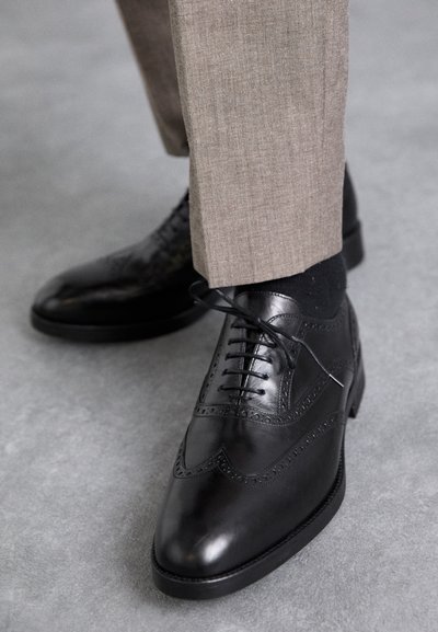 Schwarze Lederschuhe mit Broguemuster, mit spitzem Zehenbereich, Schnürdesign und glatter Textur; kombiniert mit hellbraunen Hosen.