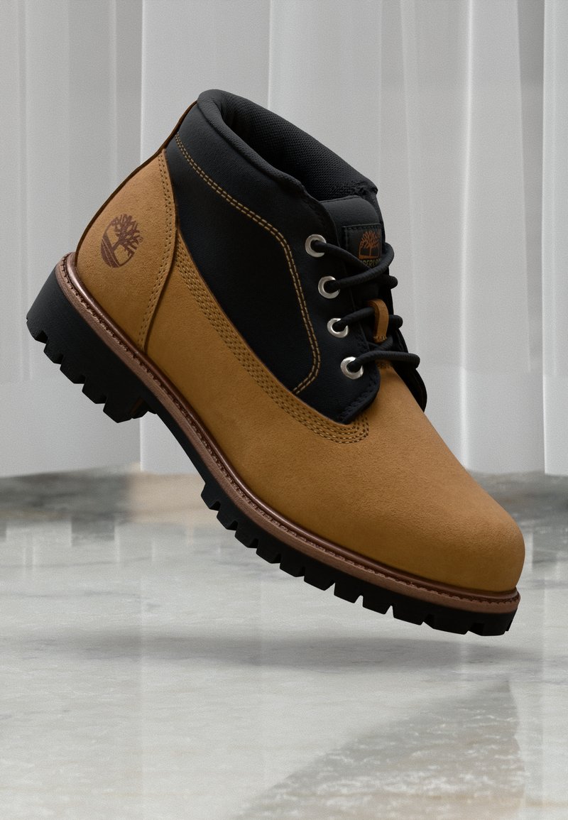 Brune semskede ankelstøvler med sort stoff-overdel, metalløpeører, gummisåle og synlige stikninger. Timberland-logo på siden.