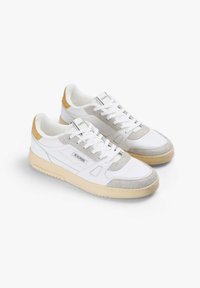 Zapatillas blancas con un acento en ante gris y amarillo, un área de cordones texturizada y una suela de goma gruesa color crema. Etiqueta de la marca visible.