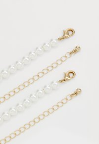 Dva náramky z perlových korálků se zlatými řetězovými články a karabinovým zapínáním na bílém pozadí.