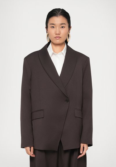 Blazer marron oversize avec un col châle, fermeture à un bouton et deux poches avant. Associé à une chemise blanche en dessous.