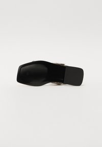 Black heeled shoe with a smooth, rectangular sole, featuring a textured surface. Accented with metallic details near the straps.
