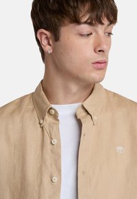 Camicia beige con bottoni realizzata in tessuto leggero, con colletto classico, bottoni frontali e un piccolo logo bianco sul lato sinistro del petto.