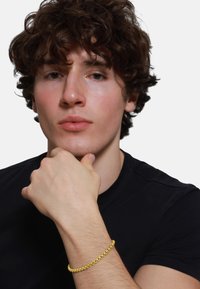 Bracelet en or présentant un design entrelacé sur un poignet, avec une finition texturée, porté par une personne en chemise noire, la main positionnée de manière réfléchie.