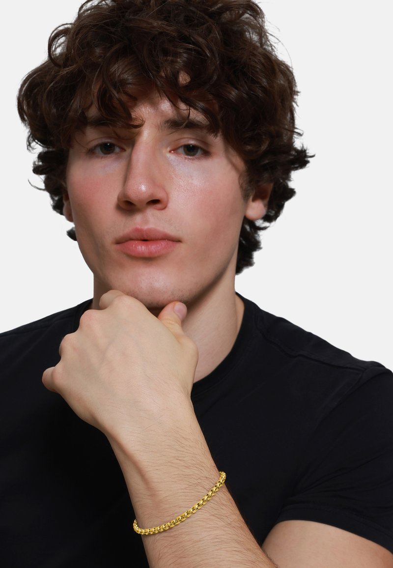 Gouden armband met een schakelpatter op een pols, met een gestructureerde afwerking, gedragen door een persoon in een zwart shirt, met de hand bedachtzaam gepositioneerd.