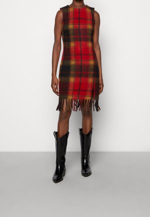 Robe sans manches à carreaux rouges et noirs avec un ourlet à franges. Fabriquée en tissu texturé, elle présente des couleurs vives et une silhouette ajustée, associée à des bottes noires.