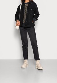 Veste en denim noir par-dessus un pull à motifs, associée à un jean foncé et des bottines beiges à lacets. Le sac est blanc, en matériau lisse.