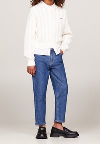 Suéter blanco de punto trenzado, superpuesto a una camisa con cuello, combinado con jeans rectos azules y mocasines negros con un acento dorado.