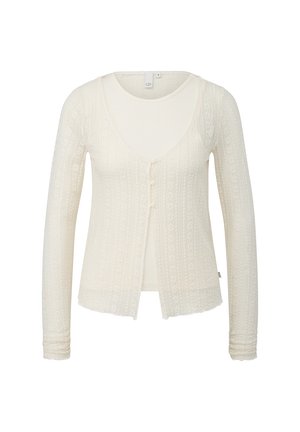 Kremowy, długi sweter na guziki z dzianiny z fakturowanym wzorem i trzema małymi guzikami z przodu, noszony na dopasowanym, bezrękawnym topie.