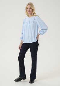 Gaiši zila blūze ar puffētu piedurknēm, kurai ir teksturēta auduma un mežģīņu akcenti uz pleciem. Apvienota ar tumšām džinsu biksēm un melnu loafers.