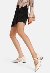 Scarpe con tacco alto brevettate con una finitura lucida e crema; due fasce decorative con fibbie; punta quadrata; tacco spesso e texturizzato.