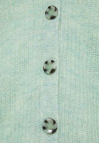 Světlý zelený pletený cardigan s čtyřmi průhlednými knoflíky s tmavými akcenty. Textura je jemná a mírně plyšová, což naznačuje teplo.