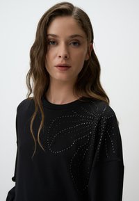 Felpa nera con un design testurizzato di strass disposti in un motivo floreale sulla spalla. Tessuto morbido con una vestibilità comoda.