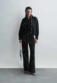 Μαύρο cropped μπουφάν με ασημένια κουμπιά, συνδυασμένο με μαύρες φαρδιές παντελόνες. Το μοντέλο κρατά μια φτιαγμένη ασημένια τσάντα με φούντες. Λείο ύφασμα, μινιμαλιστικός σχεδιασμός.