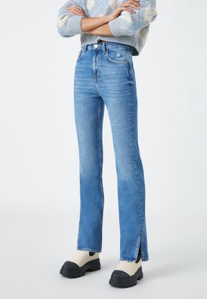 Person iført blå jeans med høj talje og slidser ved anklerne, en blå mønstret sweater og hvide sko med tyk sål og sorte tæer.