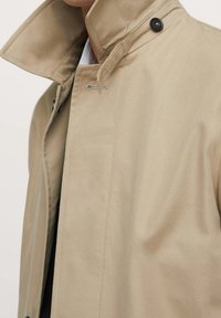 Trench-coat beige en tissu lisse, doté d'un col haut, d'une fermeture à double bouton et de détails de couture subtils.