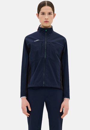 Vrouw die een marineblauwe jas met rits en bijpassende broek draagt, staand met ontspannen armen tegen een effen lichte achtergrond.