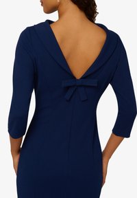 Granatowa sukienka z dekoltem w kształcie litery V i rękawami 3/4, z kokardą na dole tyłu dekoltu oraz dopasowanym fasonem.