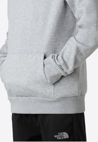 Hanorac gri deschis cu textură moale, manșete și tiv striate, buzunar tip cangur și logo în partea de jos, stânga.