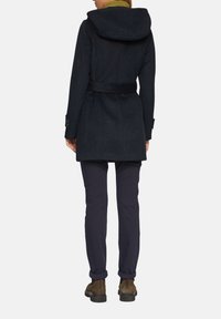 Manteau en laine bleu foncé avec capuche, taille ceinturée et poignets boutonnés, associé à un pantalon sombre et des bottines brunes. Texture lisse.