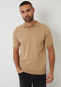 Pletené polo tričko v světle béžové barvě s krátkými rukávy a ve výstřihu do V. Obsahuje žebrované manžety a lem, se malou logem na boku.
