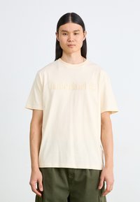 Krämfärgad bomull T-shirt med en präglad "Timberland"-logotyp på bröstet. Designen inkluderar rund hals och korta ärmar.