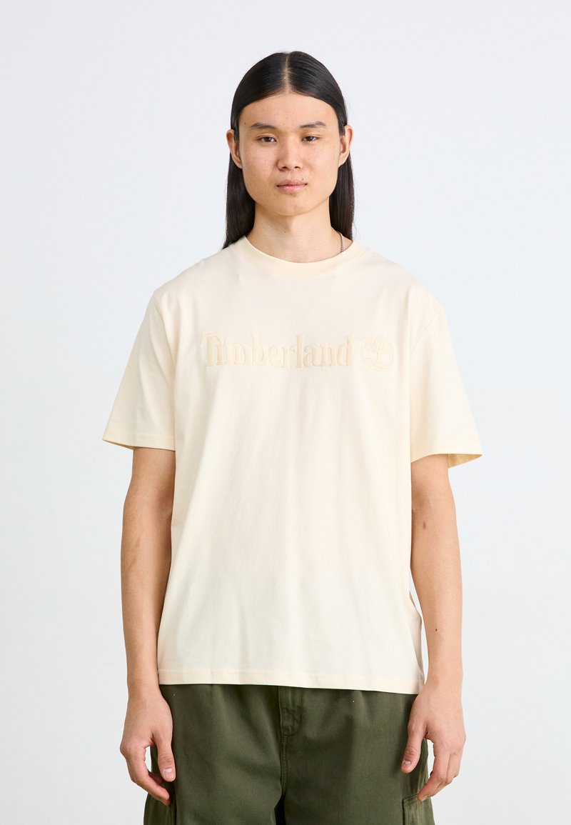 Krämfärgad bomull T-shirt med en präglad "Timberland"-logotyp på bröstet. Designen inkluderar rund hals och korta ärmar.