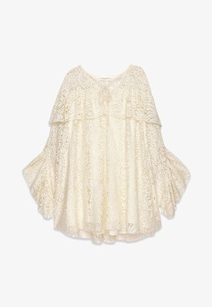 Blouse en dentelle crème avec un design superposé, des manches larges et un lien au niveau du col. Présente des motifs floraux intriqués sur l'ensemble.