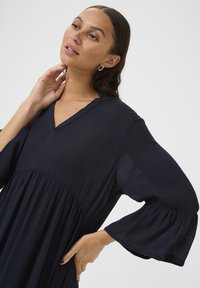 Rochie bleu marin dintr-un material ușor și texturat, cu decolteu în V, mâneci lungi tip clopot și un bust plisat.