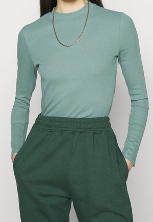 Persoon draagt een tealkleurige top met lange mouwen, ingestopt in donkergroene broek met elastische taille, handen in de zakken en een dunne gouden ketting om de hals.