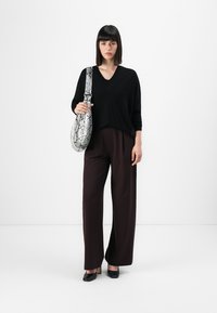 Zwarte v-hals trui, losse pasvorm, gecombineerd met bruine wijde broek. Houdt een grijze tas met slangenprint vast. Minimalistische achtergrond.