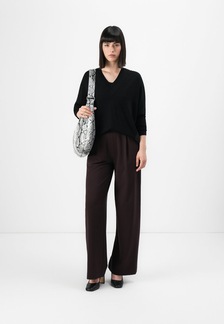 Pull noir en V, coupe ample, associé à un pantalon large marron. Tient un sac à main en imitation serpent gris. Fond minimaliste.