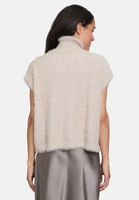 Eine von hinten sichtbare Frau trägt eine ärmellose, beigefarbene, flauschige Weste mit hohem Kragen und einen grauen Satinrock.