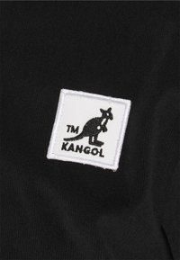 Tela negra con textura tejida que presenta un parche rectangular blanco con un logotipo de canguro y "TM KANGOL" en negro.