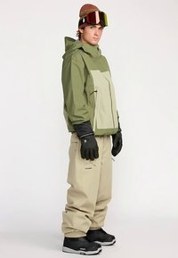 Veste de ski verte et beige avec capuche, gants noirs, pantalon de neige beige et bottes noires. Dispose d'une poche zippée et d'une coupe ample.