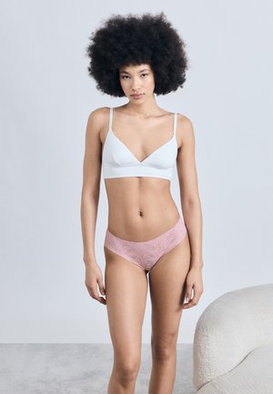 Νεαρή γυναίκα με μεγάλα σγουρά μαλλιά φοράει λευκό σουτιέν μπραλέ και ροζ δαντελένιο εσώρουχο, όρθια δίπλα σε μια καμπύλη μπεζ καρέκλα με φόντο έναν απλό τοίχο.