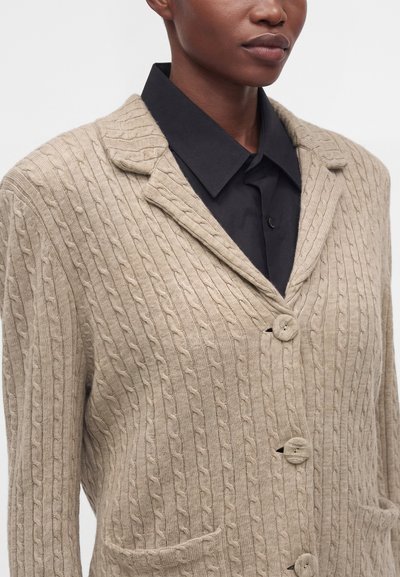 Gros plan d'une personne portant un cardigan beige en maille torsadée avec des boutons sur une chemise noire à col, sur un fond uni.