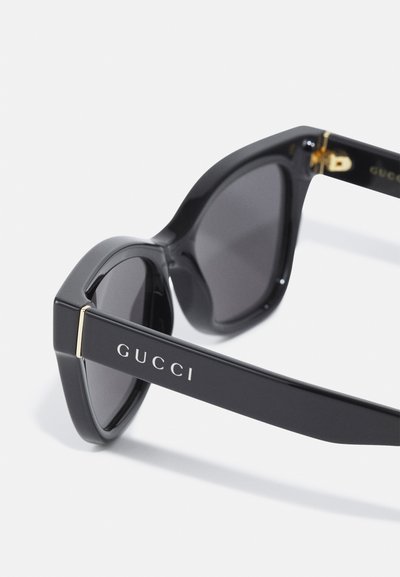 Sorte solbriller med vinklede rammer og mørke linser. Har gullhardware og "GUCCI"-logoen på brillestangen.
