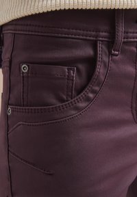 Burgunderfarvede denim jeans har en frontlomme med vertikal syning, metalknapdetalje og en glat tekstur, der fremhæver det tætsiddende design.