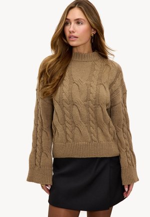 Femme aux longs cheveux ondulés portant un pull en maille torsadée beige et une jupe noire, regardant sur le côté sur un fond uni.