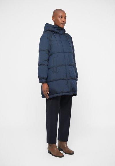 Navy blue puffer coat with a hood, featuring a quilted design, front pockets, and a relaxed fit, paired with dark trousers and brown ankle boots.