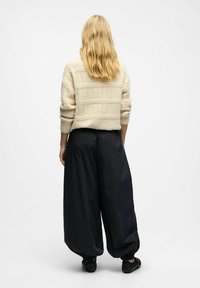 Frau mit langen blonden Haaren, die einen cremefarbenen Kabelstrickpullover, lockere schwarze Hosen mit elastischen Bündchen und schwarze Schuhe trägt, steht mit dem Rücken zum Betrachter.
