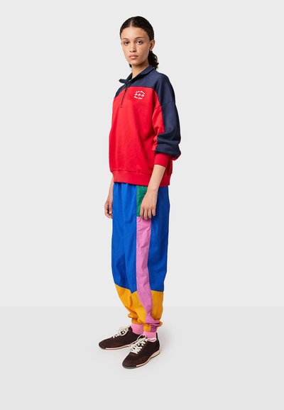 Rode en marineblauwe ritsvest met een logo, gecombineerd met meerkleurige joggingsbroek met blauwe, roze, groene en gele secties. Zwarte sneakers.
