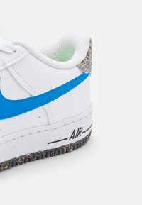 Białe sneakersy z niebieskim logo Swoosh, teksturowanym szarym obcasem i czarnym napisem "AIR" na białej podeszwie, z wielokolorową, plamkowaną podeszwą środkową.
